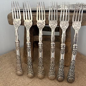 May include: A set of six silver-toned metal forks featuring intricate floral engravings on the tines and ornate, embossed scrollwork patterns along the handles. The forks are arranged in a row, showcasing a consistent design with detailed metalwork.