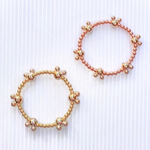 Può includere: Due braccialetti dorati con piccole perle a forma di fiore. I braccialetti sono su uno sfondo bianco.
