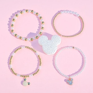 May include: Four beaded bracelets with gold accents and white pearls, along with a white Mickey Mouse-shaped decoration. One bracelet has a Mickey Mouse charm with blue accents. The items are arranged on a pink surface.
