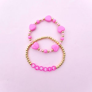 May include: Two pink beaded bracelets with gold beads. The top bracelet has pink heart-shaped beads and the bottom bracelet has white letters spelling out "BARBIE".