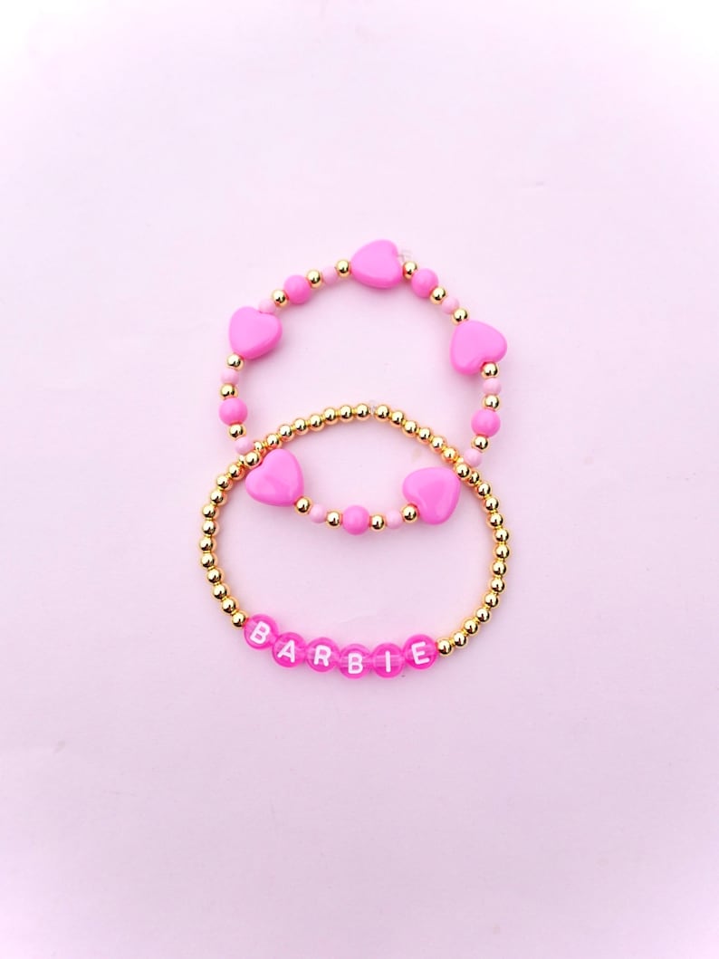 May include: Two pink beaded bracelets with gold beads. The top bracelet has pink heart-shaped beads and the bottom bracelet has white letters spelling out "BARBIE".