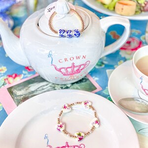 May include: Two bracelets with floral designs. The first bracelet is gold with blue and white floral beads. The second bracelet is gold with white beads and pink and white floral beads. Both bracelets are on white plates with a crown design.