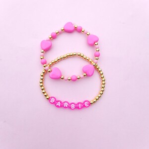 May include: Two pink beaded bracelets with gold accents. The top bracelet has pink heart-shaped beads and the bottom bracelet has pink beads with white letters spelling out "BARBIE".