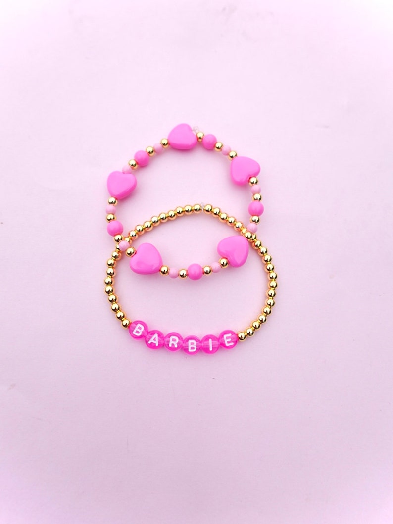 May include: Two pink beaded bracelets with gold accents. The top bracelet has pink heart-shaped beads and the bottom bracelet has pink beads with white letters spelling out "BARBIE".