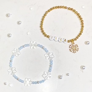 Könnte beinhalten: Zwei Armbänder mit Schneeflockenanhängern. Ein Armband ist golden mit dem Namen "Elsa", der in weißen Perlen geschrieben ist. Das andere Armband ist blau mit durchsichtigen Schneeflockenperlen.