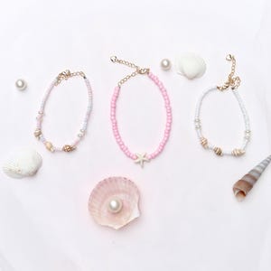 Op de afbeelding: Drie kralen enkelbandjes in pastelkleuren met schelp- en zeesteraccenten. De enkelbandjes worden gepresenteerd met schelpen en parels op een witte achtergrond, wat een strandthema oproept. Elk enkelbandje heeft een goudkleurige ketting.