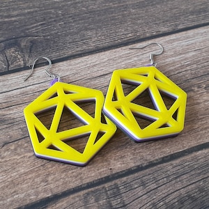 May include: A pair of geometric earrings with a yellow, white, purple, and black color scheme. The earrings are made of layered material and have a silver hook. The earrings are displayed on a wooden surface.