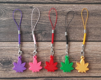 May include: Five colourful phone charms with a leaf design. The charms are purple, pink, red, green, and yellow. Each charm is attached to a braided cord.