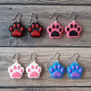 May include: Six pairs of dangle earrings featuring a paw print design. The earrings are made of black, pink, white, and blue plastic. The paw prints are 3D and have a textured surface.