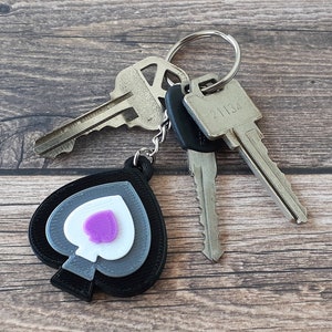 May include: A black, gray, and white spade-shaped keychain with a purple heart in the center. The keychain is attached to a set of keys with a silver key ring.