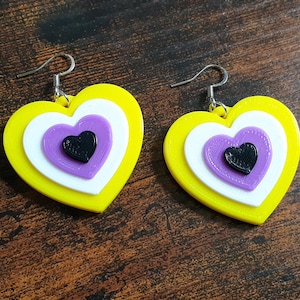 Peut inclure: Une paire de boucles d'oreilles en forme de cœur jaunes, blanches et violettes avec un cœur noir au centre. Les boucles d'oreilles sont en plastique imprimé en 3D.