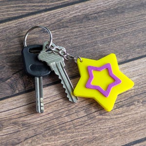 Puede incluir: Un juego de llaves con un llavero en forma de estrella amarilla con un contorno de estrella púrpura. Las llaves están en un llavero plateado, sobre un fondo de madera.