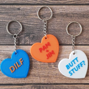 May include: Three heart-shaped keychains with different phrases. The first keychain is blue with the word "DILF" in orange letters. The second keychain is orange with the words "Pam & Jim" in red letters. The third keychain is white with the words "Butt Stuff?" in blue letters.