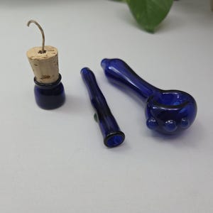 Peut inclure: Un ensemble de trois objets en verre bleu cobalt comprenant une petite bouteille avec un bouchon en liège et une mèche, un tube en verre fin et une pipe en verre arrondie avec trois petites bosses décoratives sur le côté du foyer.