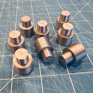 以下が含まれることがあります： 8つの小さな円筒形の金属オブジェクトのコレクション。各ピースは、テクスチャ加工されたリブ付きベースと滑らかな平らな上面を備えています。オブジェクトは、青と緑のグリッド表面に配置されています。
