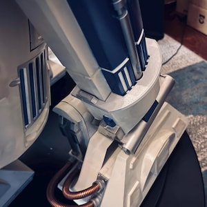 Puede incluir: Primer plano de un modelo detallado en blanco roto y azul marino. El modelo presenta detalles plateados metálicos, incluyendo tubos y conectores. El diseño tiene una estética futurista, con un enfoque en formas geométricas y componentes mecánicos.