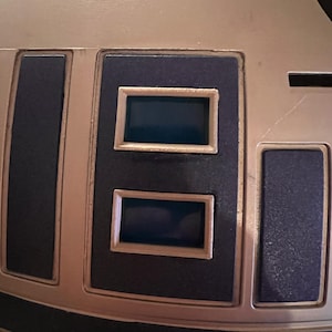 Puede incluir: Primer plano de una superficie metálica, posiblemente robótica, con paneles rectangulares azul oscuro. Se ven dos aberturas rectangulares, enmarcadas por un borde beige claro. El diseño sugiere un tema futurista o tecnológico.