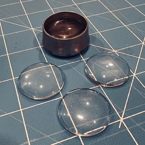May include: A small, dark metal container with liquid inside, alongside three clear, circular glass cabochons. The items are on a blue cutting mat with white grid lines. The cabochons are approximately 4 cm in diameter.