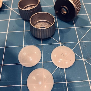 Könnte beinhalten: Eine Ansammlung von silberfarbenen, geriffelten Knöpfen und drei durchscheinenden, runden, weißen Kunststoffteilen ist auf einer blauen Rasteroberfläche angeordnet. Die Knöpfe haben eine zylindrische Form mit gerillter Außenseite.