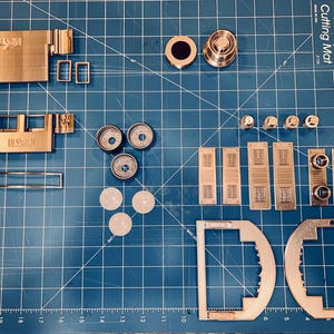 以下が含まれることがあります： 青いカッティングマットの上に配置された、機械装置と思われる金属部品の詰め合わせ。長方形や円形の部品、ネジなどのハードウェアが含まれています。部品は様々な形状とサイズです。