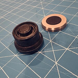 Puede incluir: Dos objetos metálicos sobre una superficie azul. Uno es un objeto cilíndrico oscuro con anillos concéntricos y una superficie texturizada. El otro es un objeto circular plateado con un centro negro. Son componentes mecánicos.