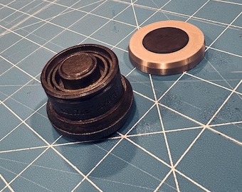 May include: Two metal objects sit on a blue surface. One is a dark, cylindrical object with concentric rings and a textured surface. The other is a silver-colored, circular object with a black center. The objects appear to be components of a larger mechanism.