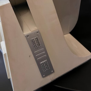 Puede incluir: Primer plano de una sección de un objeto color crema con una placa de metal plateado adjunta. La placa tiene forma rectangular con líneas verticales grabadas y una barra horizontal. El objeto tiene un diseño curvo.