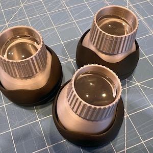 May include: Three magnifying glasses. Each has a silver-coloured, ridged top and a clear lens. The base is a rounded, black shape. They are arranged on a blue grid surface.