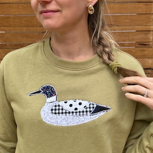 Könnte beinhalten: Ein olivgrünes Sweatshirt mit Rundhalsausschnitt und einer zentralen Stoffapplikation in Form einer Ente. Die Ente besteht aus verschiedenen gemusterten Stoffstücken, darunter florale Muster, schwarze und weiße Punkte sowie Vichy-Karos.