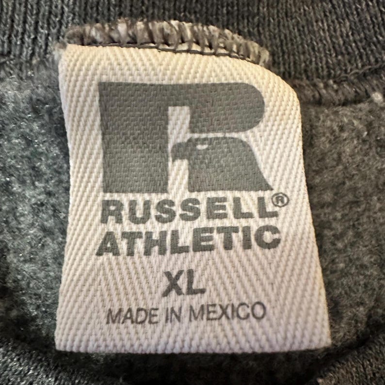 K&ouml;nnte beinhalten: Nahaufnahme eines grau-wei&szlig;en Russell Athletic-Etiketts. Das Etikett zeigt das Logo der Marke, den Text "RUSSELL ATHLETIC" und die Gr&ouml;&szlig;e "XL". Auf dem Etikett steht auch "MADE IN MEXICO".