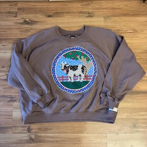 May include: A brown Gap sweatshirt with a circular patch featuring a black and white cow in a field with a tree and red apples. The patch is surrounded by a blue border with white dots and a red and green border.
