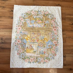 May include: A vintage, cream-colored fabric featuring a charming illustration of mice in a cozy cottage setting. The scene is framed by a floral border of pink and blue flowers. The mice are depicted in various activities, such as reading and having tea.
