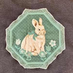 K&ouml;nnte beinhalten: Ein hellbraunes Kaninchen mit einer t&uuml;rkisfarbenen Schleife sitzt auf einem t&uuml;rkisfarbenen Hintergrund mit floralen Akzenten. Das Bild wird von einem gewellten Rand umrahmt. Das Design befindet sich auf einem dunkelgrauen Stoff.