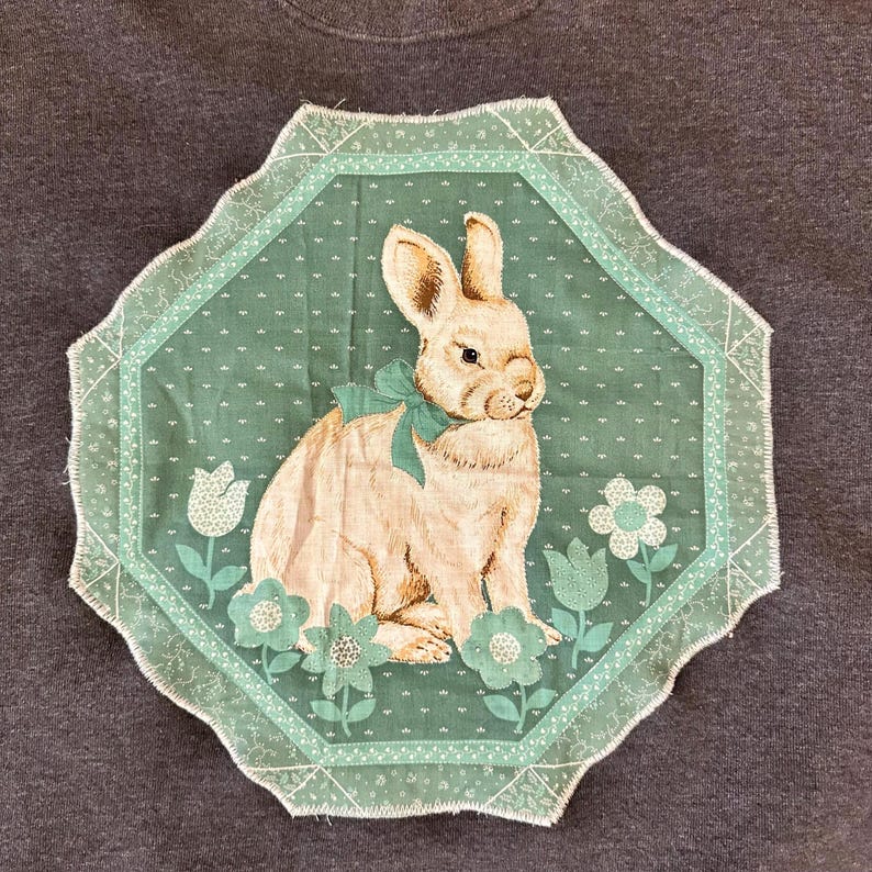 K&ouml;nnte beinhalten: Ein hellbraunes Kaninchen mit einer t&uuml;rkisfarbenen Schleife sitzt auf einem t&uuml;rkisfarbenen Hintergrund mit floralen Akzenten. Das Bild wird von einem gewellten Rand umrahmt. Das Design befindet sich auf einem dunkelgrauen Stoff.
