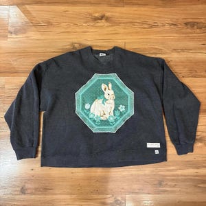 K&ouml;nnte beinhalten: Dunkelgraues Sweatshirt mit einem wei&szlig;en Kaninchen-Design in einem t&uuml;rkis-wei&szlig;en achteckigen Rahmen. Das Kaninchen hat eine t&uuml;rkisfarbene Schleife und ist von Blumen umgeben. Das Sweatshirt hat lange &Auml;rmel und einen Rundhalsausschnitt, geeignet f&uuml;r Freizeitkleidung.