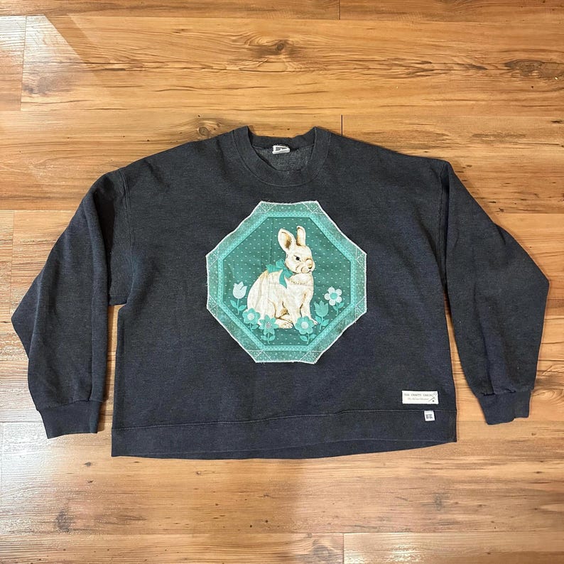 K&ouml;nnte beinhalten: Dunkelgraues Sweatshirt mit einem wei&szlig;en Kaninchen-Design in einem t&uuml;rkis-wei&szlig;en achteckigen Rahmen. Das Kaninchen hat eine t&uuml;rkisfarbene Schleife und ist von Blumen umgeben. Das Sweatshirt hat lange &Auml;rmel und einen Rundhalsausschnitt, geeignet f&uuml;r Freizeitkleidung.