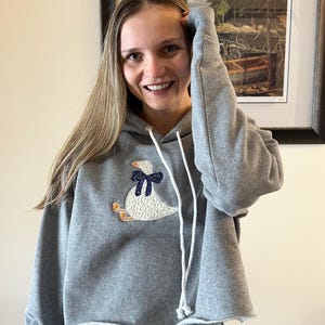 Könnte beinhalten: Ein grauer, gecroppter Kapuzenpullover mit weißem Kordelzug und einem dekorativen Gänse-Applikation. Die Gans hat einen weißen Körper, eine blaue Schleife und orangefarbene Füße. Der Hoodie wird mit Jeans kombiniert.