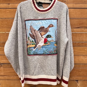 Peut inclure: Un pull à manches longues gris chiné avec un grand écusson rectangulaire sur le devant représentant deux canards colverts volant au-dessus d'un lac bleu. Le pull présente un col rond côtelé et des poignets avec des rayures crème et rouge foncé.