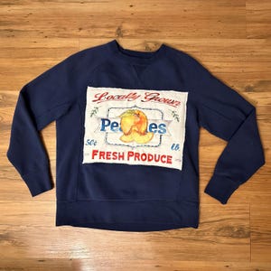以下が含まれることがあります： 紺色のスウェットシャツ。桃のイラストと「Locally Grown Peaches Fresh Produce」の文字が入ったパッチが付いています。パッチはヴィンテージ風の手縫い風で、水色の縁取りがあります。