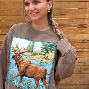 May include: A brown crewneck sweatshirt featuring a large, square, centred graphic print of a brown moose standing in a blue river surrounded by green pine trees, tall grasses, and cattails. The sweatshirt has long sleeves with ribbed cuffs.