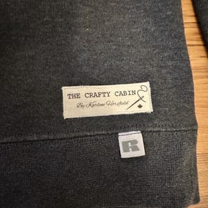 K&ouml;nnte beinhalten: Dunkelgraues Sweatshirt mit einem wei&szlig;en Etikett mit der Aufschrift "THE CRAFTY CABIN By Karlene Horsfield". Ein kleines wei&szlig;es Etikett mit einem grauen "R" ist unten angen&auml;ht. Das Sweatshirt ist aus einem weichen, strukturierten Stoff.