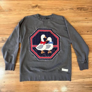 May include: Gray sweatshirt with a large, octagonal patch featuring two white ducks with red and blue accents. The patch is bordered by a red and white pattern. The sweatshirt has a crew neck and long sleeves.