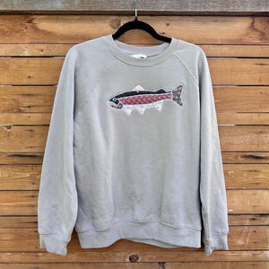 May include: A light grey crewneck sweatshirt featuring a central appliqué of a fish. The fish is constructed from multiple fabric patches with floral and geometric patterns in shades of red, white, black, and blue, outlined with white stitching.