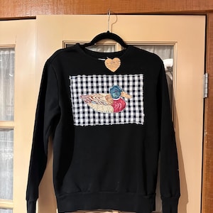 Peut inclure: Un sweat-shirt noir à manches longues avec un empiècement rectangulaire à motif vichy bleu marine et blanc. L'empiècement présente une illustration de canard colvert coloré. Une étiquette en forme de cœur avec du texte est fixée au col.