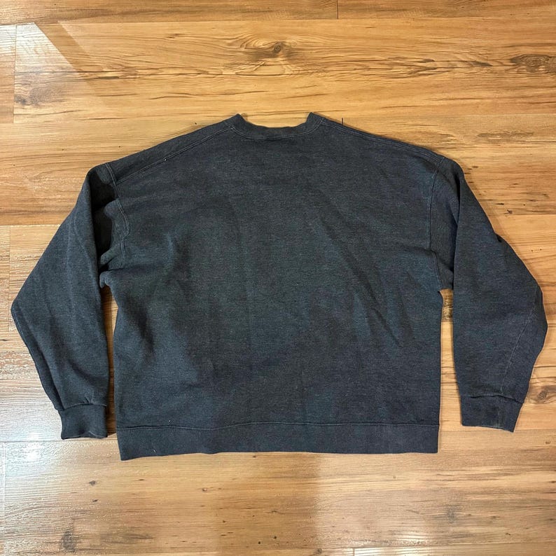 K&ouml;nnte beinhalten: Ein dunkelgraues Sweatshirt mit Rundhalsausschnitt und langen &Auml;rmeln. Das Sweatshirt besteht aus einem weichen Material und hat gerippte B&uuml;ndchen und Saum. Das Kleidungsst&uuml;ck liegt flach auf einer Holzoberfl&auml;che.