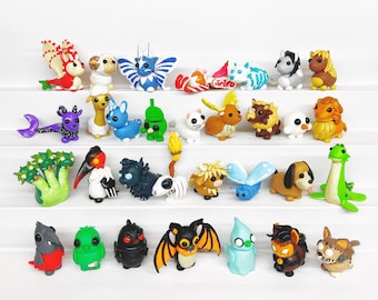 Peut inclure: Une collection de 30 figurines en pâte polymère faites à la main et de couleurs vives. Les figurines représentent divers animaux, créatures mythologiques et objets. Elles sont disposées en rangées sur une surface blanche.