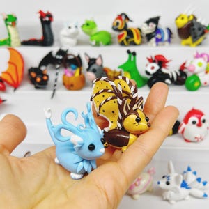May include: Two small, hand-crafted clay figurines. One is a blue dragon with white accents and the other is a brown and yellow fox with white stripes. The figurines are sitting on a hand.