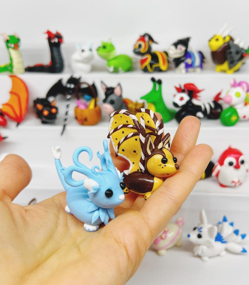 May include: Two small, hand-crafted clay figurines. One is a blue dragon with white accents and the other is a brown and yellow fox with white stripes. The figurines are sitting on a hand.