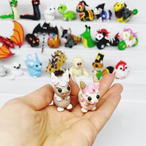May include: Two handmade polymer clay figurines of cute animals. The figurine on the left is a white and brown cat with a pink nose and brown eyes. The figurine on the right is a pink and white pig with a pink nose and black eyes. Both figurines are holding a small, white, heart-shaped object.