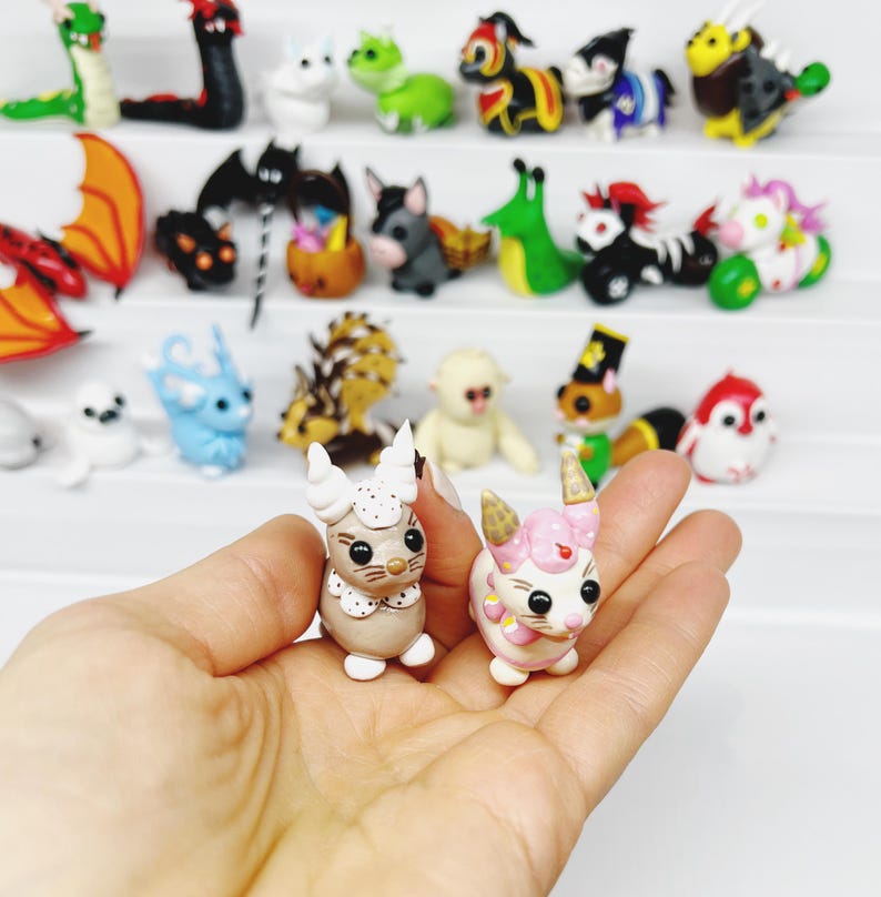 May include: Two handmade polymer clay figurines of cute animals. The figurine on the left is a white and brown cat with a pink nose and brown eyes. The figurine on the right is a pink and white pig with a pink nose and black eyes. Both figurines are holding a small, white, heart-shaped object.